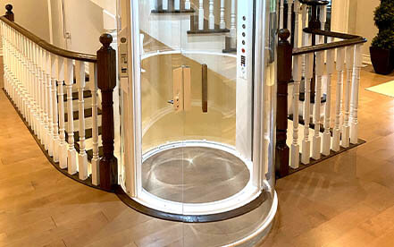 Glass home elevator at the base of a traditional staircase with wooden railings