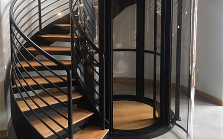 Glass residential elevator positioned beside a curved modern staircase