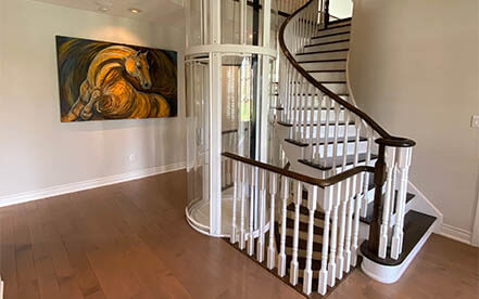 Glass residential elevator installed beside a curved staircase in a traditional home.