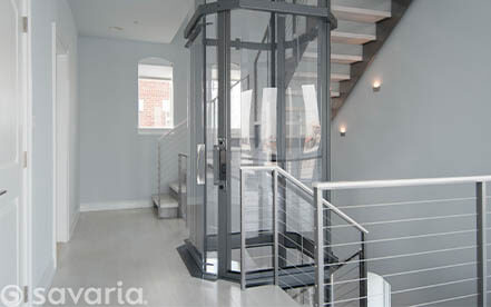 Glass residential elevator inside a modern hallway with a metal staircase.